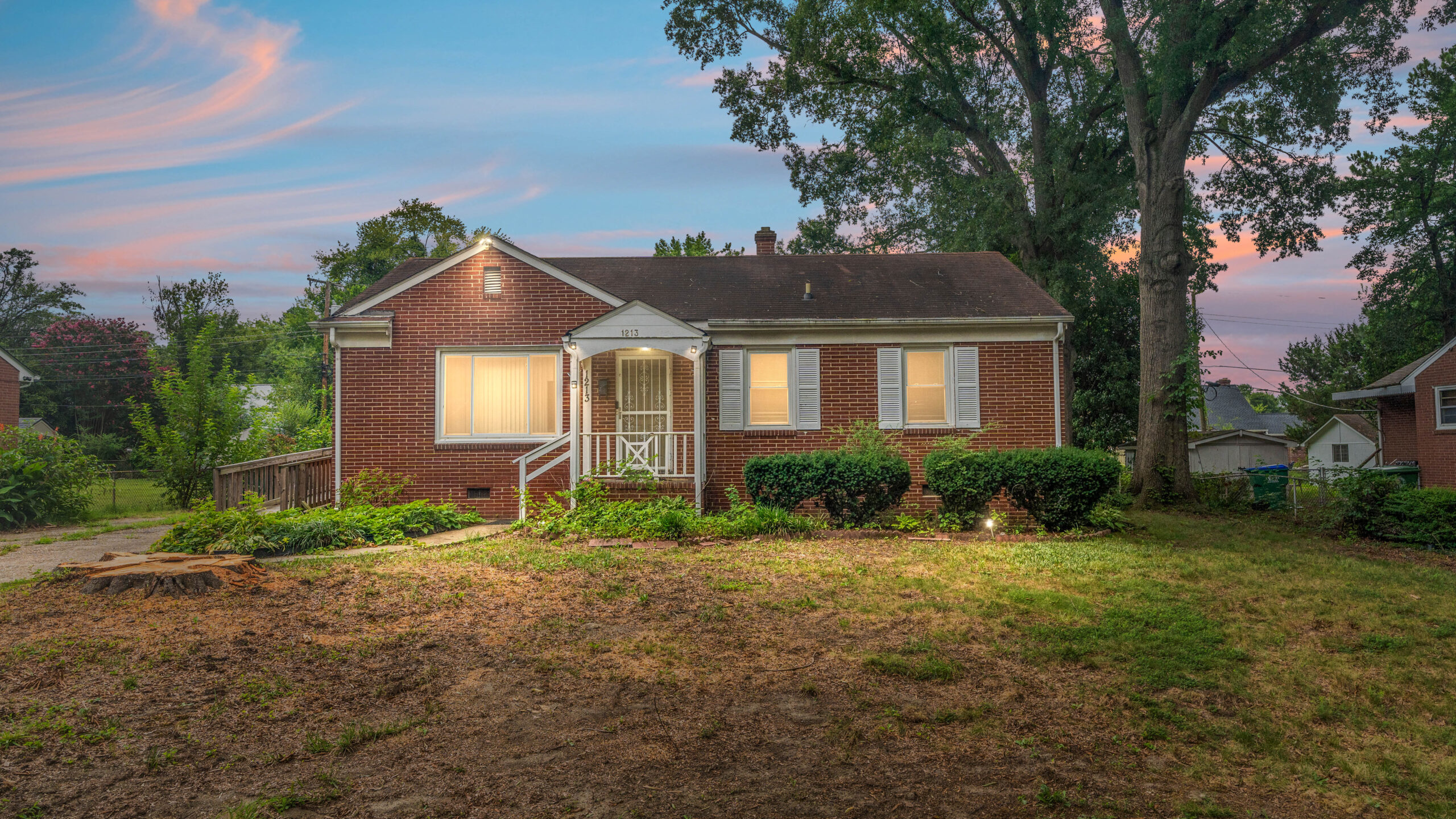 Brick, ranch-style home with a large tree to the right and bushes out front